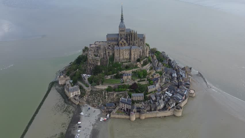 Aerial view of the legendary medieval island commune of Le Mont-Saint-Michel and its iconic Gothic abbey soaring above the tidal flats in Normandy, France. Cinematic drone shot showcasing historic ramparts, winding medieval village streets, soaring spire, and surrounding bay on a sunny day.