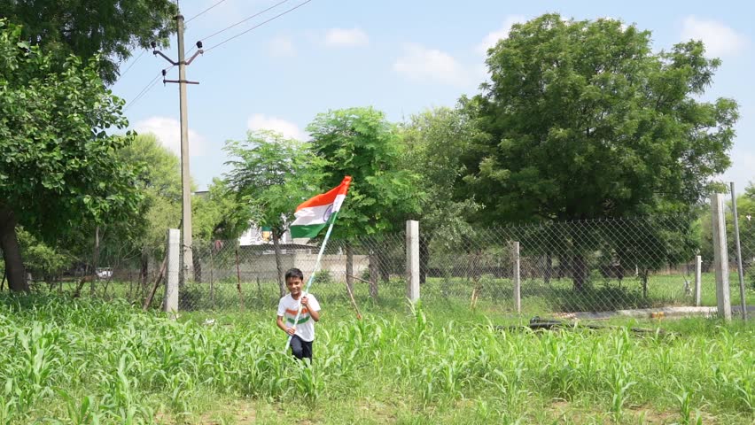 Indian village kids wearing national flag logo design printed t shirt and holding, waving or running with Tricolour with greenery in the background, celebrating Independence or Republic day