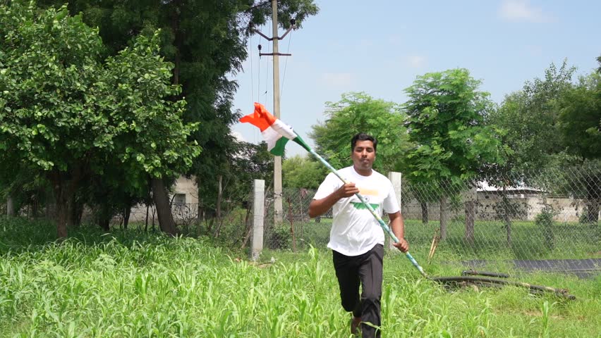Indian village kids wearing national flag logo design printed t shirt and holding, waving or running with Tricolour with greenery in the background, celebrating Independence or Republic day