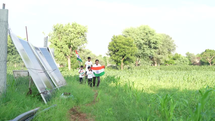 Indian village kids wearing national flag logo design printed t shirt and holding, waving or running with Tricolour with greenery in the background, celebrating Independence or Republic day