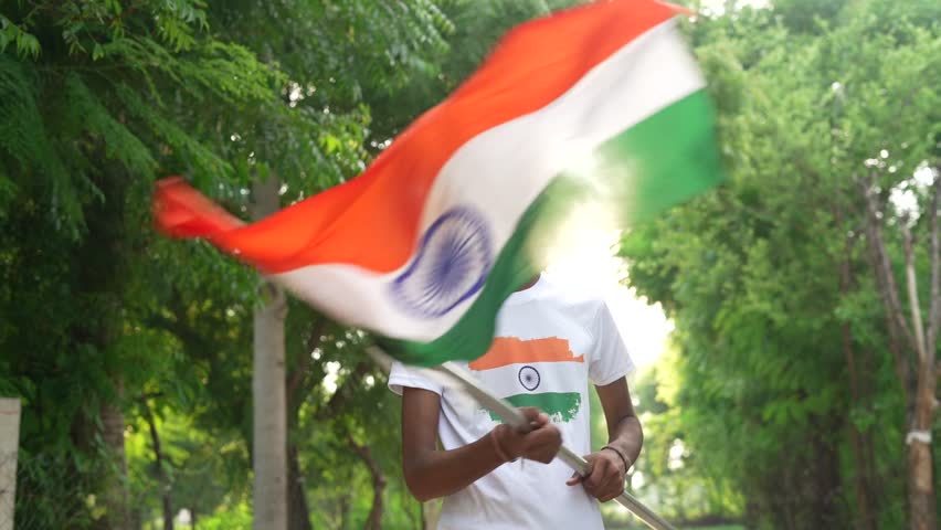 Indian village kids wearing national flag logo design printed t shirt and holding, waving or running with Tricolour with greenery in the background, celebrating Independence or Republic day
