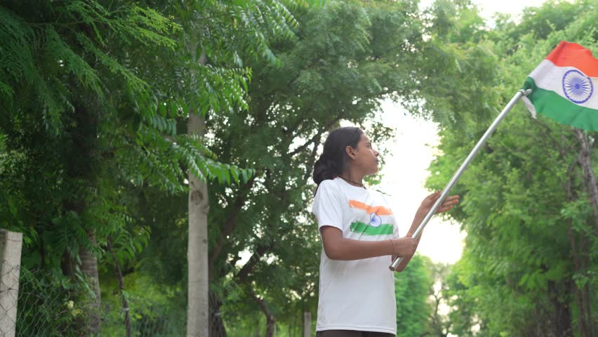 Indian village kids wearing national flag logo design printed t shirt and holding, waving or running with Tricolour with greenery in the background, celebrating Independence or Republic day