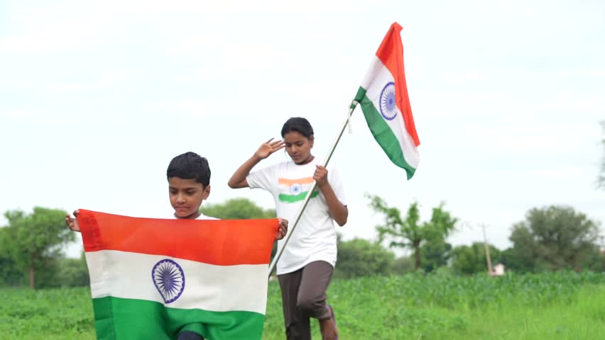 Indian village kids wearing national flag logo design printed t shirt and holding, waving or running with Tricolour with greenery in the background, celebrating Independence or Republic day