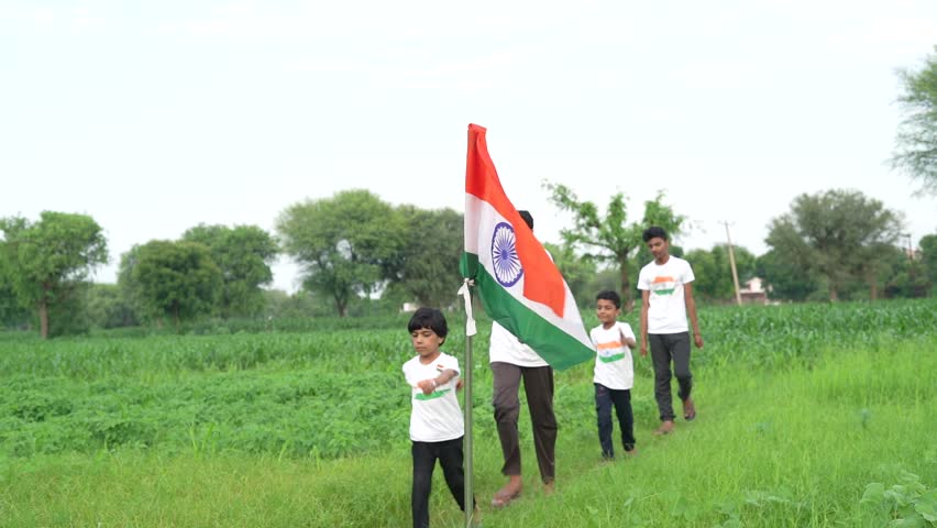 Indian village kids wearing national flag logo design printed t shirt and holding, waving or running with Tricolour with greenery in the background, celebrating Independence or Republic day
