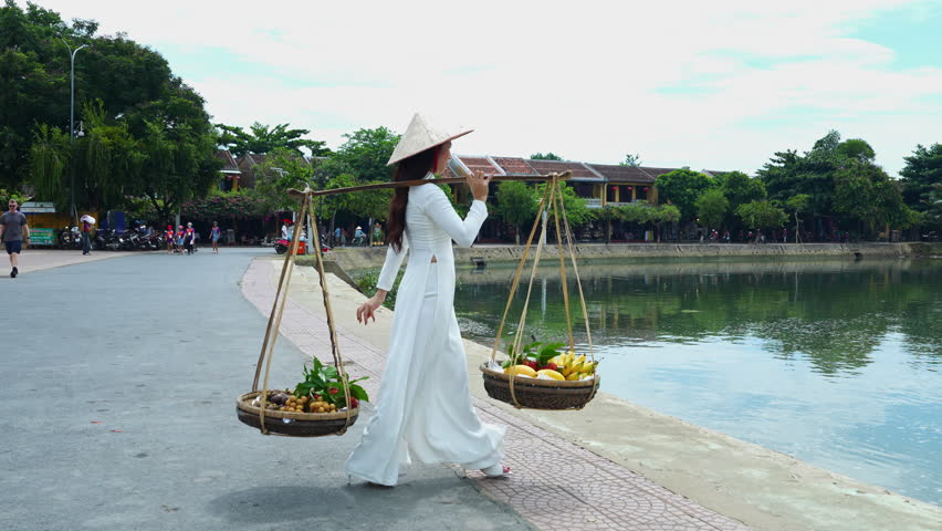 Asian Woman In Traditional Vietnamese Conical Hat Carrying Yoke With Whicker Baskets With Bananas And mango. At Hoi An, Vietnam