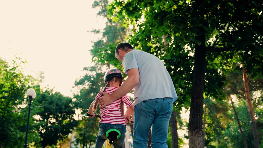 Child cyclist with Father, nature. Family weekend, Father teaches kid daughter to ride bike in park. Happy family, little girl in helmet together with her dad learns to ride bicycle outdoors in summer