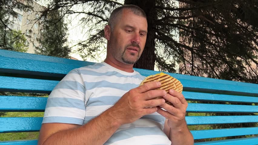 man eats Gyros on a walk: a grilled bun stuffed with potatoes, other vegetables, sauce and meat.