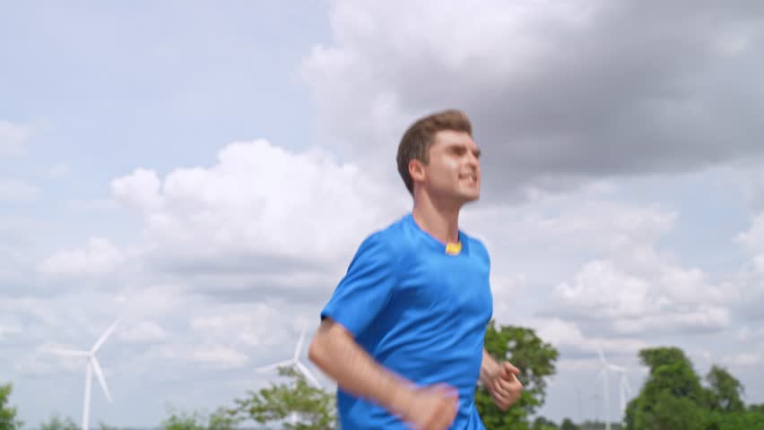 Caucasian active sportsman running exercise at the wind turbine field. Attractive athlete runner in sportswear doing jogging workout outdoors for health care in rural area at windmill power farm.