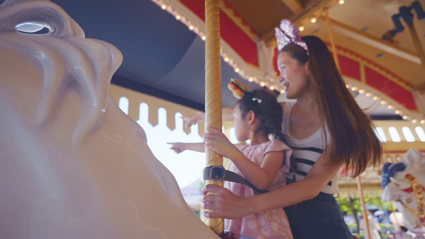 Asian mother and adorable kid daughter riding carousel at theme park. Happy family, attractive woman and little girl feeling happy and relax while traveling for holiday vacation trip at amusement park
