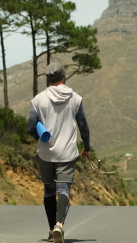 a sporty man in shorts and sports leggings walks along a road in a park near high mountains and holds a yoga mat and a jump rope in his hands. stylish sportsman man walking after jogging with yoga mat
