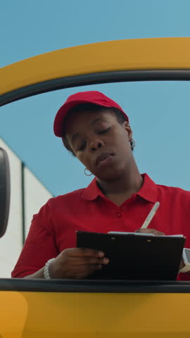 Vertical shot of young black woman in red cap and T-shirt signing delivery form when leaving car outdoors