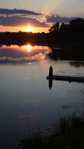 Amazing sunset over Khorol River in Myrhorod, Ukraine. Vertical video, 31 sec. Peaceful sunset over a serene lake with a silhouette of a person standing on a dock. Captures the tranquility of nature