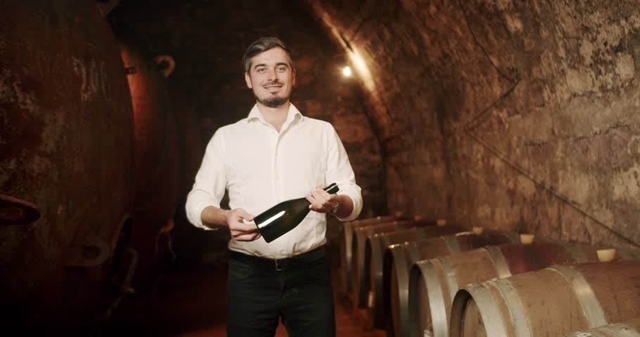 Man show the wine bottle in a factory, old cellar with many wooden barrels. Sommelier admiring the wine in the old cellar. Male winemaker is tasting a red wine in a wine cellar. Winemaking concept.