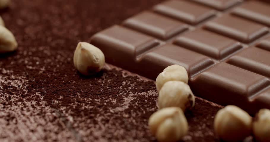 A close-up of a chocolate bar with scattered hazelnuts on a richly textured cocoa powder surface. Perfect for food, dessert, and confectionery themes. Delicious Milk Chocolate Bar with Hazelnuts