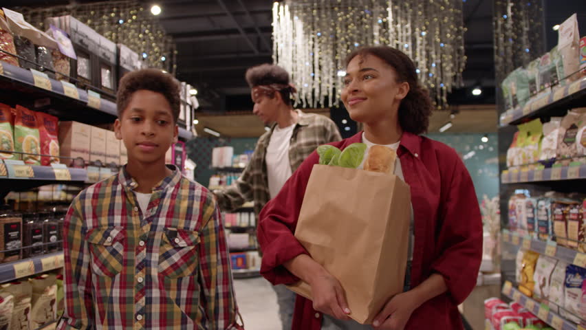 Positive relatives walking around store with filled shelves and making grocery shopping together. Mom, dad and son buying food at supermarket with big variety of product brands and household goods.