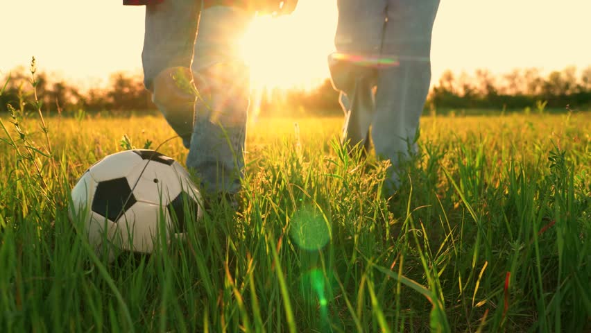Legs of happy children kids kicking ball, playing football in field at golden sunset. Cheerful playful children kids playing football, kicking soccer ball into camera. Funny joyful game at meadow.