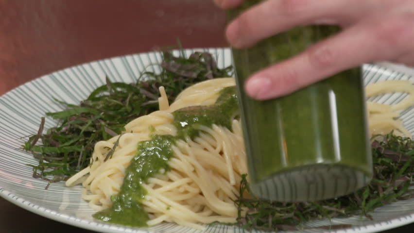 Person pouring green sauce on spaghetti with seaweed stems