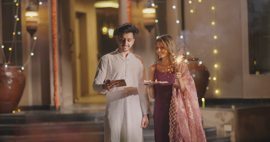 Indian Hindu happy gen z couple pair standing hold mobile phone take cute funny pose selfie outdoor home. Smiling young adult husband wife enjoy Diwali festival celebrate festive season outside house