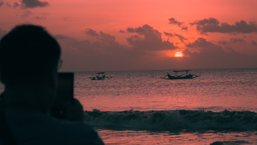 Silhouetted Man Takes Photo of Majestic Burning Sunset over Sea at Jimbaran Beach Landscape with Fishing Boats Moored Near the Shore