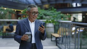 Handsome Asian Gentleman Walking from Coffee Break to Workplace and Using Phone. Smiling Manager in Glasses Is Texting with Colleagues. People and Technology Concept - Powered by Shutterstock - Get 15% off with code: PIKWIZARD15