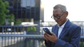 Smiling Asian Businessman Using Cell Phone While Standing on the Business Centre Background. Busy Senior Man Checking Email, Chatting, Browsing Social Media. People and Technology Concept - Powered by Shutterstock - Get 15% off with code: PIKWIZARD15