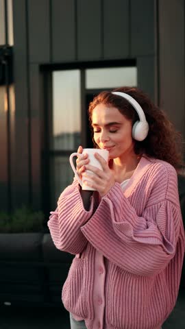 A Young Curly-Haired Woman Wearing Headphones Approaches the Railing on the Terrace and Takes a Sip of Tea or Coffee. The Girl Admires the View of the City During the Sunrise. Vertical Shot