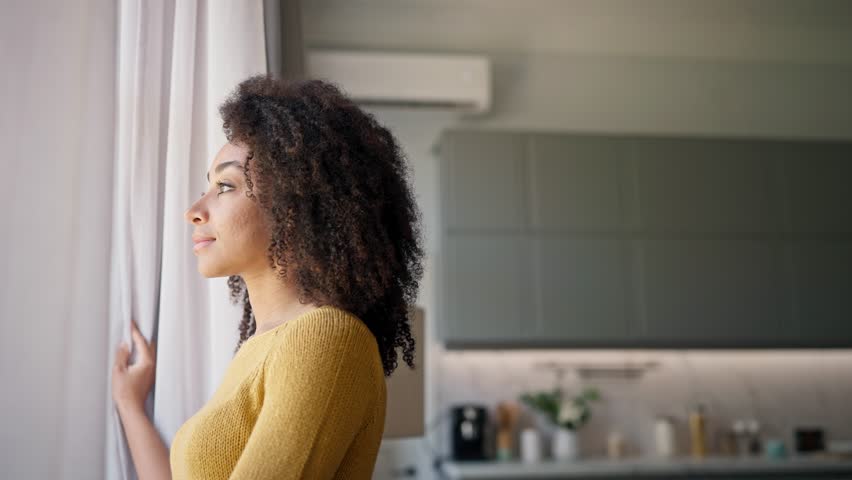 Portrait of African American woman with curly hair standing near window and holding curtains with one hand. Happy female smiling with joy while glancing at camera. In background visible modern kitchen
