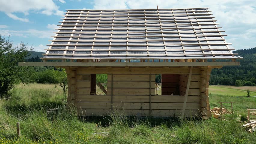 Wooden house under construction, made of logs and located in beautiful landscape during a beautiful summer day, surrounded by lush greenery, grass and trees under a blue sky