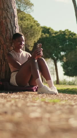 Vertical video, Young woman in casual clothes using her smartphone outdoors, chatting on social media, talking online with friends, union with nature and mental health concept
