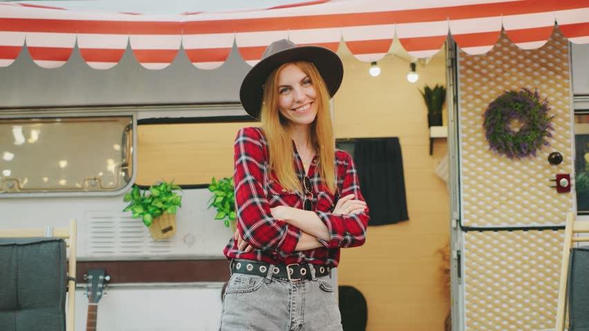 A young woman with red hair is wearing a stylish hat and a casual shirt while standing in front of a camper van. The scene suggests that she is on a travel adventure. The camper and the natural