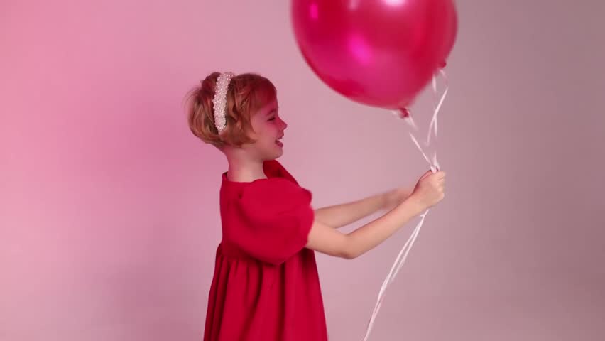 A young girl is holding four red balloons in her hand. She is sitting on the ground and looking up at the balloons. Concept of innocence and joy, as the girl is surrounded by the colorful balloons