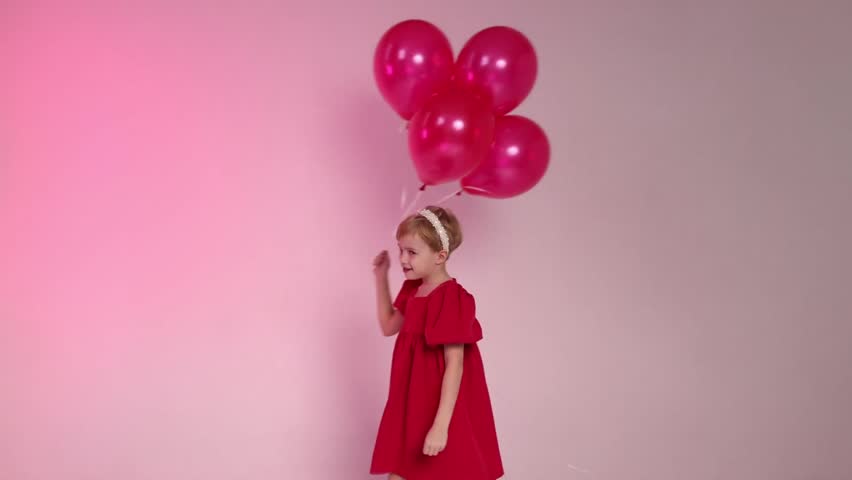A young girl is holding four red balloons in her hand. She is sitting on the ground and looking up at the balloons. Concept of innocence and joy, as the girl is surrounded by the colorful balloons