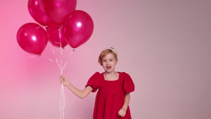 A young girl is holding four red balloons in her hand. She is sitting on the ground and looking up at the balloons. Concept of innocence and joy, as the girl is surrounded by the colorful balloons