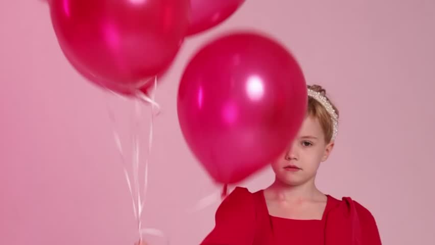 A young girl is holding four red balloons in her hand. She is sitting on the ground and looking up at the balloons. Concept of innocence and joy, as the girl is surrounded by the colorful balloons