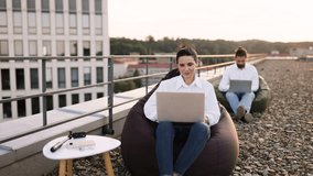 Two business people working outdoors on rooftop with laptops. Casual work environment with bean bag chairs and scenic city view. Focus on relaxation and productivity. - Powered by Shutterstock - Get 15% off with code: PIKWIZARD15