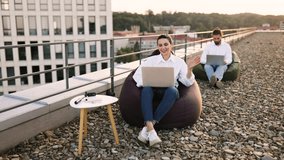 Two professionals working on laptops seated on bean bags on rooftop. Relaxed and productive outdoor workspace with cityscape view. Concept of remote work team collaboration and modern work environment - Powered by Shutterstock - Get 15% off with code: PIKWIZARD15