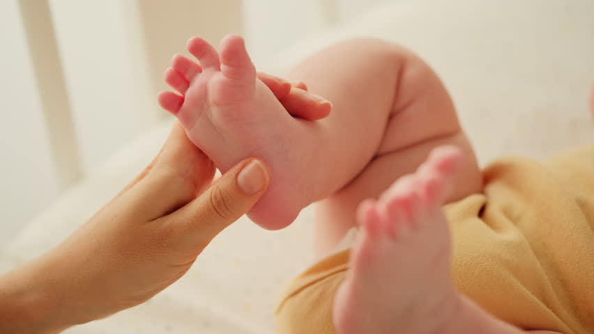 Mother holding in hands foot of newborn baby. Close up of woman gently stroking, touching tiny feet of baby boy. Family care, happy childhood and motherhood.