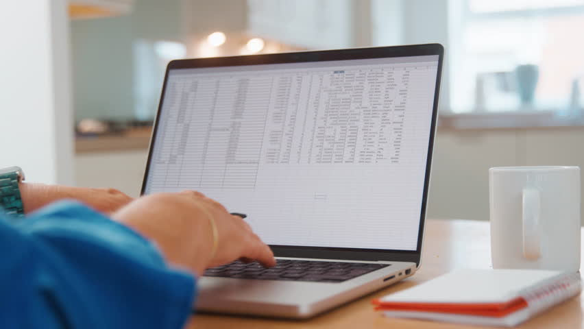 A person types on a laptop with spreadsheets open at a home office.