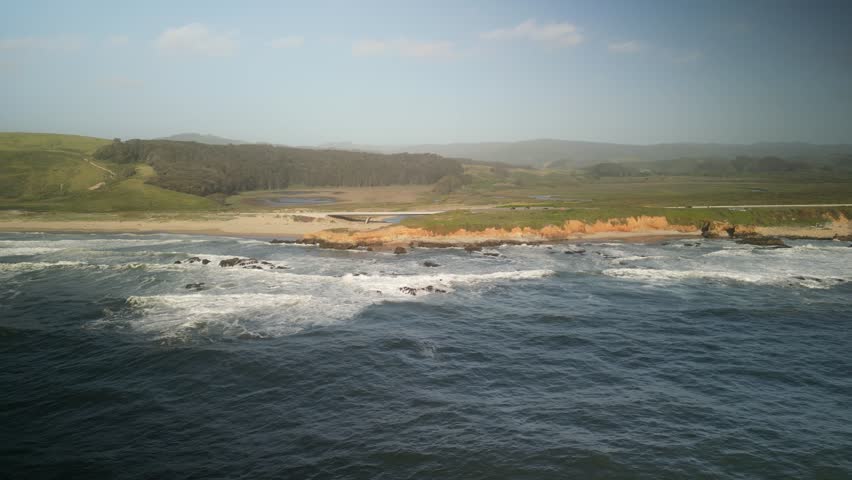 Aerial drone footage over Pescadero State Beach from the Peninsula of Northern California. The coastline features rugged cliffs, sandy shores, and crashing waves, all bathed in golden sunlight.