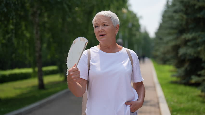 Suffering from heat of mature woman fanning herself in park by strong summer heat wave. Unhappy gray-haired older female fanning with fan suffering heat stroke on summer in city park, slow motion.
