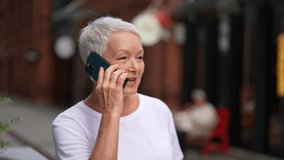 Closeup face of happy European gray-haired older female talking on phone outdoors. Smiling senior adult woman using cellphone making call standing on city street outside. Shooting in slow motion. - Powered by Shutterstock - Get 15% off with code: PIKWIZARD15