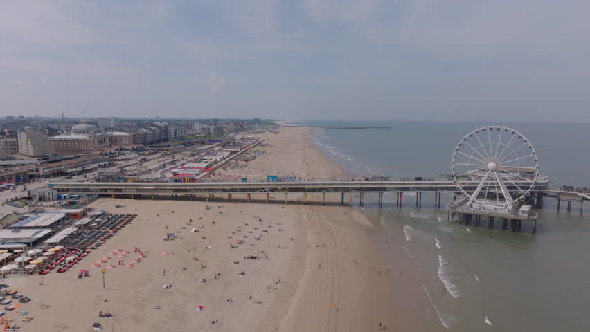 Aerial view of The Hague beach with pier and ferris wheel on a sunny day. Urban landscape and coastline in the background, beachgoers enjoying the ocean waves