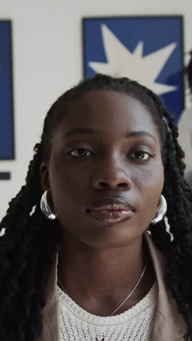 Vertical portrait shot of young Black woman with dreadlocks sitting in modern art gallery and posing for camera