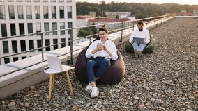 People working remotely on rooftop using laptops and bean bags. Man focused on laptop, woman talking on phone. Relaxed outdoor work environment with cityscape background - Powered by Shutterstock - Get 15% off with code: PIKWIZARD15