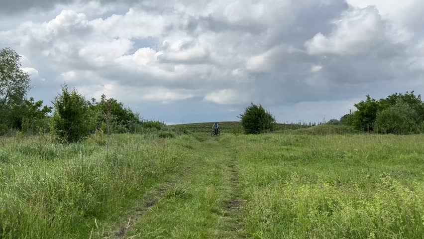 Mountain biker rides through lush green countryside path surrounded by trees and fields under a cloudy sky. Serene rural landscape for outdoor adventure