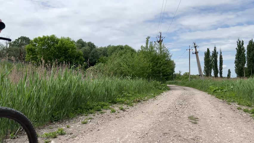 Cyclist leisurely rides on a rural dirt path, surrounded by lush green trees and tall grass under a partly cloudy sky