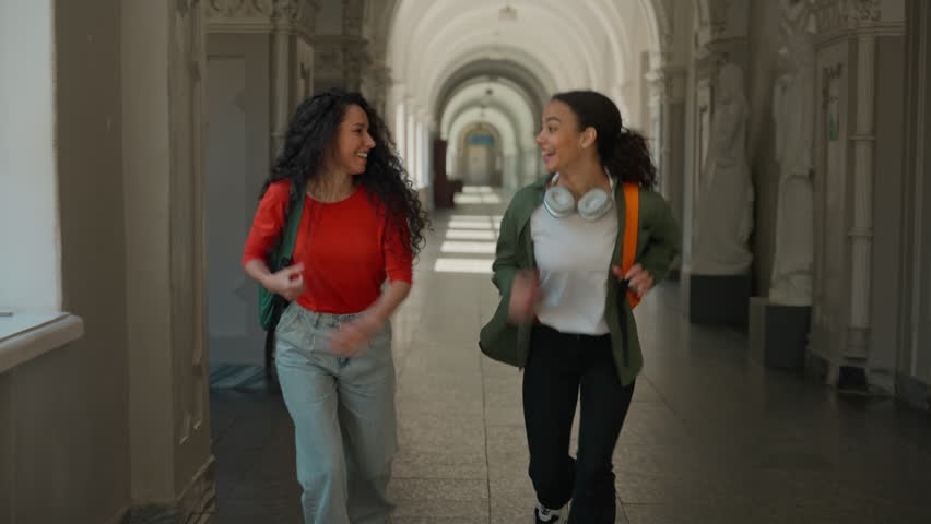 Two happy active running young students girls smiling laugh females women friends Hispanic Latina multiracial ladies run hurry rush to course lecture class in academy college university late studying