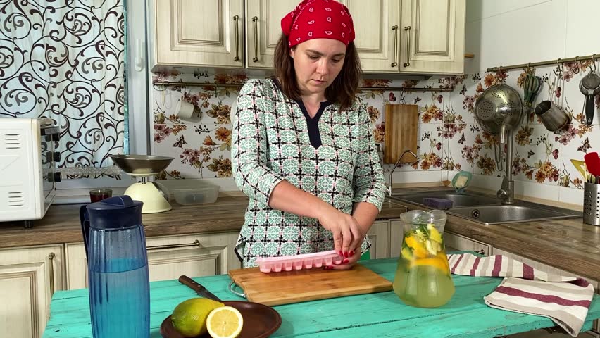 A young woman in an apron prepares ingredients for making a refreshing lemonade in home kitchen. She combines citrus fruits, mint, ice to create a cool, invigorating drink. Homemade summer beverages