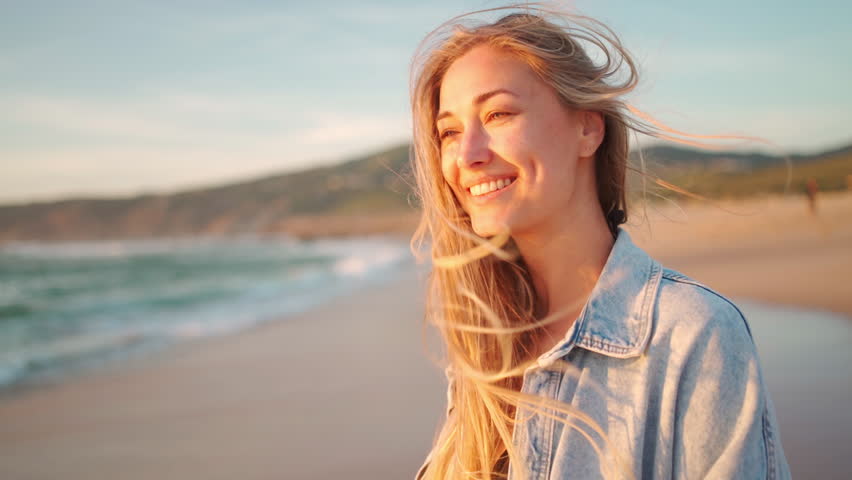 Happy woman with long blond hair at windy beach during sunny day. Cheerful female tourist is looking at camera and smiling during vacation. 