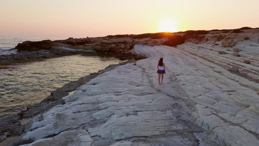 Beautiful young girl walking along the rocky shore at the sunset. Coral bay, Cyprus